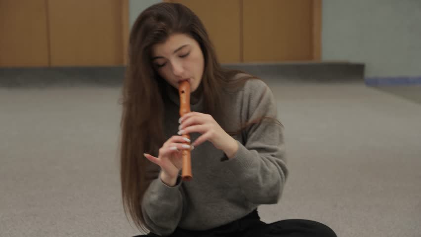 Talented young woman with long hair sitting on the floor and playing a classical melody on a wooden soprano recorder with concentration