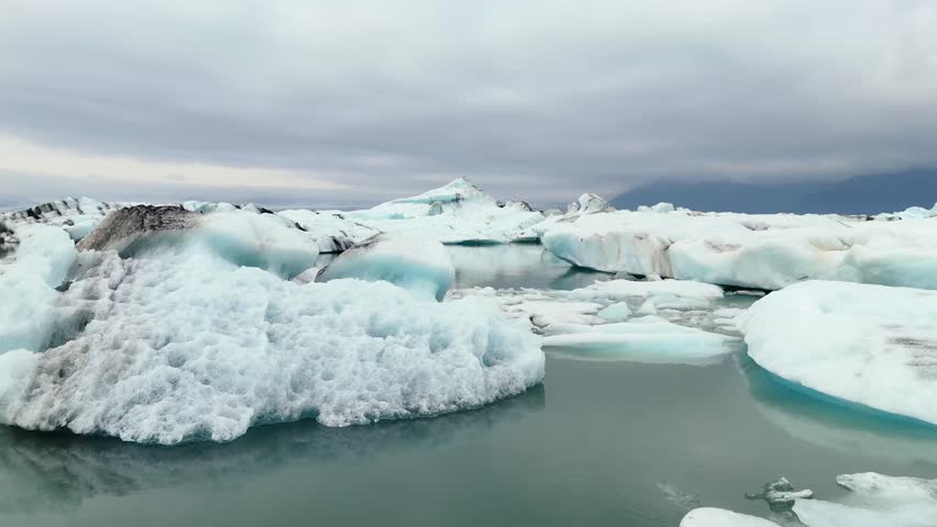 Aerial footage of icebergs scattered across the calm glacier lagoon at Jokulsarlon, Iceland, with cold blue water and a remote Nordic atmosphere.