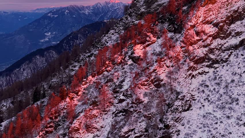 Drone footage of a snow-covered mountain side at dusk. Crimson light illuminates rocks and sparse trees, overlooking a deep valley and distant peaks.