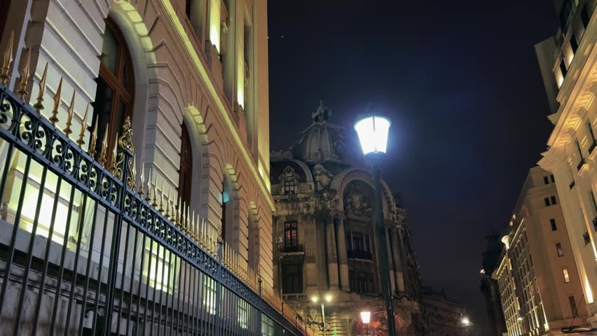 A low-angle night view of the historical Burse Chamber of Commerce building in Bucharest, Romania, illuminated by streetlights, with an ornate fence in the foreground.