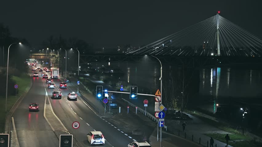 Vehicles navigate a busy road at night in Warsaw, Poland, with the illuminated ÅšwiÄ™tokrzyski Bridge and Vistula River in the background. Reflects urban life and movement.