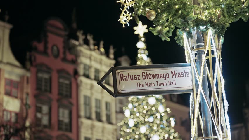 Direction sign to Gdansk Main Town Hall, adorned with festive Christmas lights and greenery, with illuminated buildings and a large Christmas tree in the blurred background at night.