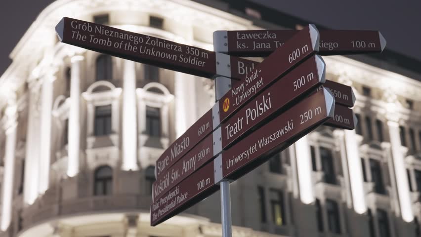 An illuminated street signpost in Warsaw, Poland, directs visitors to historical landmarks like the Old Town and the Tomb of the Unknown Soldier, showcasing the city