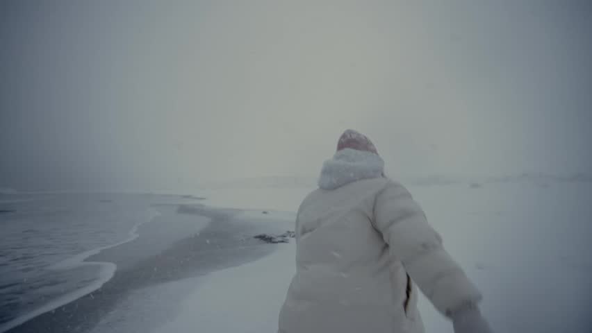 Person in red hat walking along snowy shoreline under overcast sky