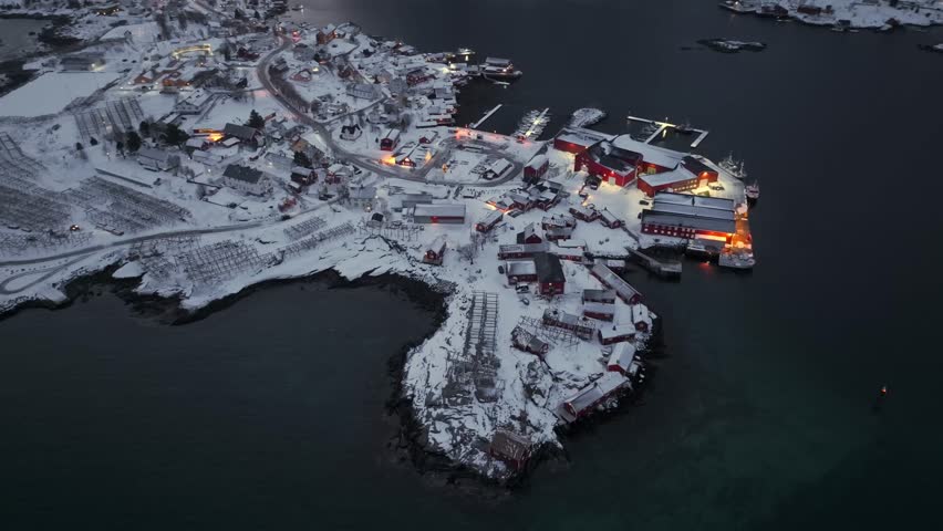 Aerial dusk view of snowy coastal village on rocky peninsula with illuminated waterfront