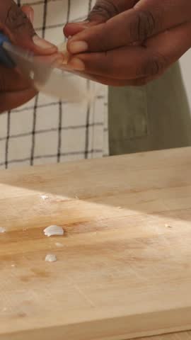 Vertical video: Peeling hands holding clove, slicing mincing garlic on board for meal, plaid apron. Knife, preparation, kitchen, closeup, culinary, countertop, vertical video