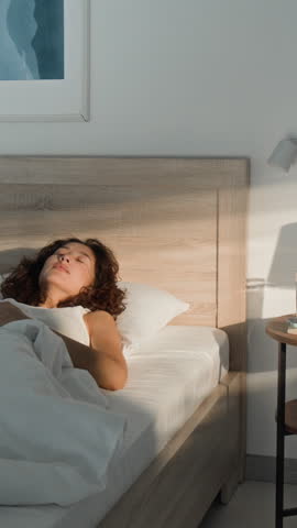 Vertical shot of Asian young adult woman waking up in bed, checking smartphone, stretching, and waving at camera. Ideal for morning routine, wellness apps, or mobile tech ads