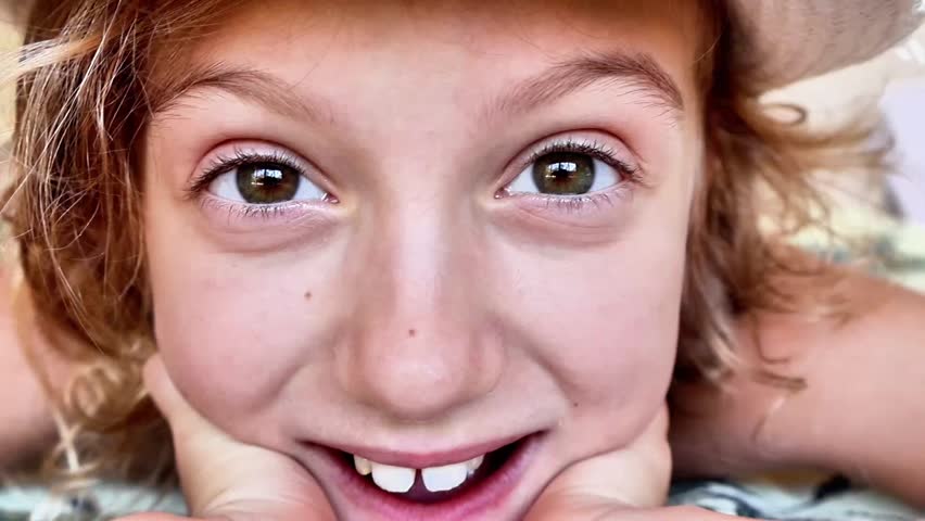 An extreme close-up portrait focusing on the face of a happy young blonde girl. She is smiling warmly at the camera with her chin resting gently on her hands. Her expressive brown eyes, light freckles, and a slight gap between her front teeth are captured with high clarity, evoking a sense of pure joy, innocence, curiosity, and authentic childhood emotion.