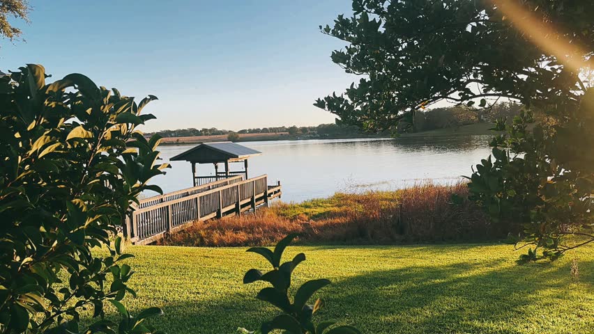 A covered wooden dock sits beside calm lake water, framed by leafy green trees, shrubs, and a bright grassy lawn. Warm sunlight enters from the upper right, adding a golden glow across the shoreline and soft water surface. The setting includes open sky, distant trees, and a quiet waterfront atmosphere.