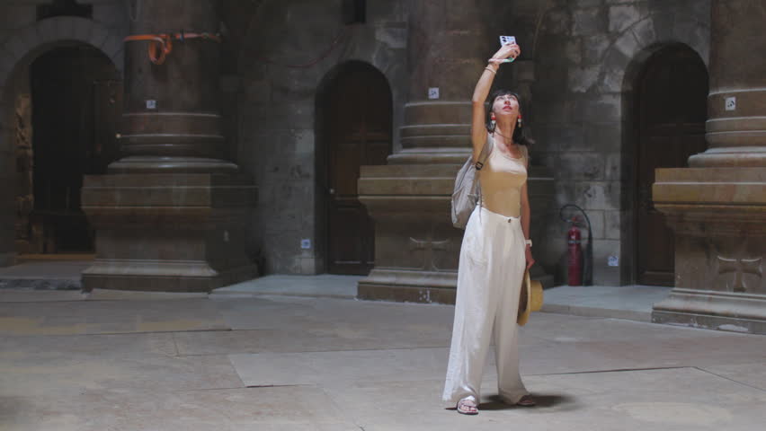 A woman wearing a flowing white dress stands in a sunlit historic plaza with large stone arches and columns. She raises a phone to her face and frames a selfie. Church of the Holy Sepulchre. Israel.