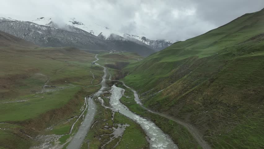 High-angle aerial view capturing a clean, rushing mountain creek flowing from a melting snow patch down a vibrant green alpine valley with rocky terrain and small wildflowers in Ushguli, Georgia
