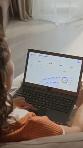 Vertical shot of young adult Caucasian woman analyzing business dashboard on laptop at home. Footage suitable for remote work, digital marketing, and e-commerce analytics promotions