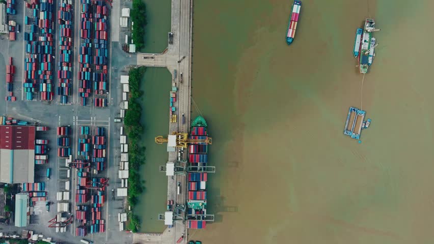 Aerial tracking shot of container ships loading and unloading at industrial port terminal along muddy river with top down Ho chi minh, Vietnam.