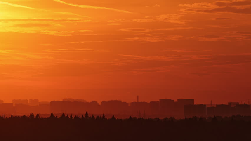 Sunset sky over city skyline with silhouette of trees, time lapse. Bright orange sun disk lowering behind clouds at dusk. Dramatic evening landscape view for weather background and travel concept.