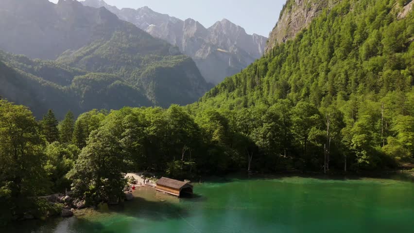 Aerial approach toward a wooden boathouse on the emerald lake shore surrounded by forest and mountains, Konigssee area, Berchtesgaden National Park, Bavaria, Germany