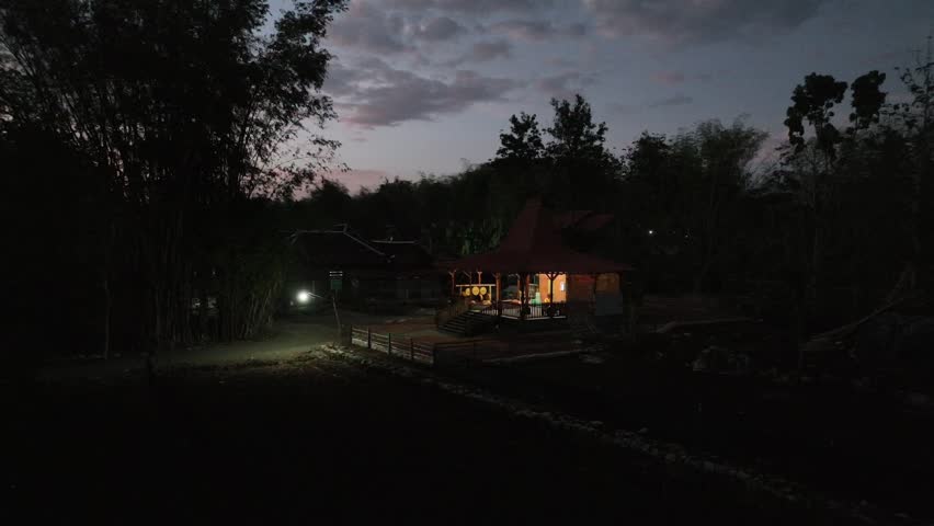 Dark forest edges frame a lone cottage lit warmly at dusk. Soft glow spills from windows, inviting calm and solitude. Silent trees stand watch as twilight deepens around the home