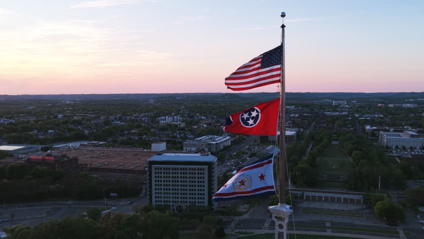 Aerial drone view of the US, Tennessee, and Nashville flags flying from the Tennessee State Capitol flagpole at dusk with the city spreading to the horizon.