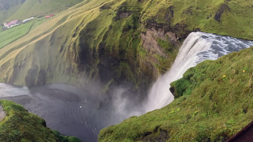 Slow motion footage of a bird flying towards and away from Skogafoss waterfall in Iceland seen from above the cliff edge. Powerful white cascade drops into a misty pool below with lush green hills