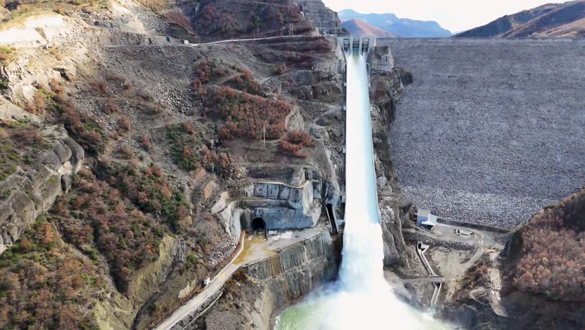 Aerial drone rising while tilting down over the Moglicë Dam spillway in Albania, capturing rushing white water roaring down the concrete channel into the mountain gorge