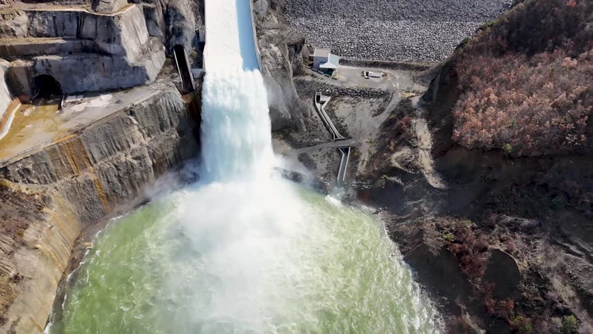 Aerial camera pulls back from a close-up of water rushing down the spillway chute to reveal the massive rock-fill embankment wall of the Moglicë dam in Albania