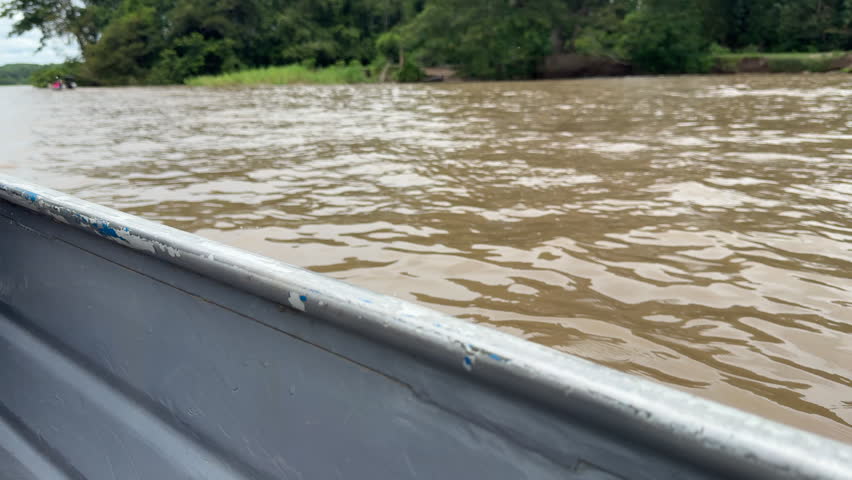 A boat travels along a muddy river, bordered by dense green jungle. Ideal for travel content or documentaries on Amazonian ecosystems.