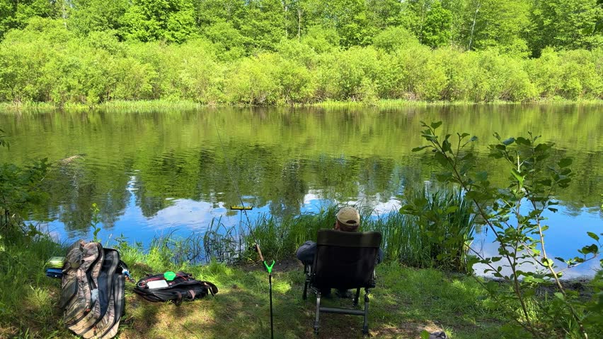 A fisherman relaxes on a chair by a calm forest lake with fishing gear, peaceful summer fishing and picturesque freshwater landscape.
