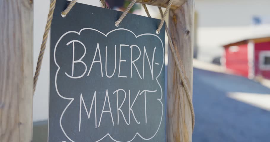 A close-up of a rustic hand-lettered chalkboard sign reading "Bauernmarkt" (farmers market) hanging from a wooden frame with rope in a rural Lower Austrian town. The shallow depth of field blurs the street and buildings behind, keeping focus on the charming artisan signage that evokes local produce, community, and authentic Austrian rural culture.