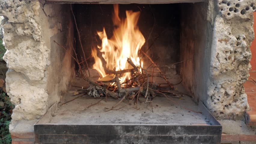 Person tending small wood fire inside rustic outdoor stone fireplace, daylight use