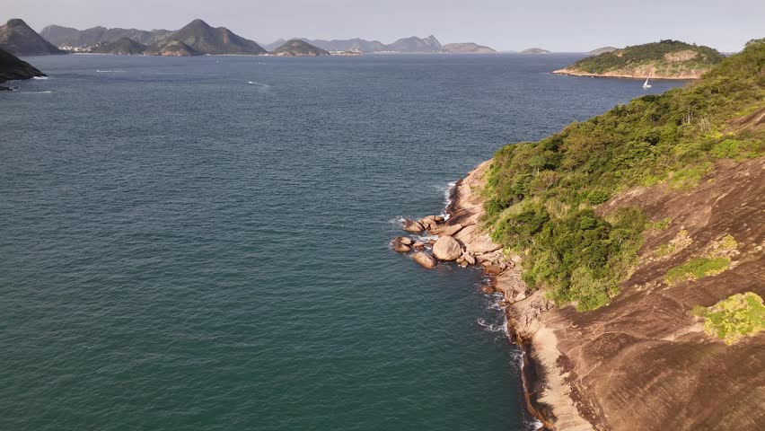 View of Guanabara Bay featuring rocky shoreline, lush green vegetation, and distant mountains under a clear sky in Brazil.