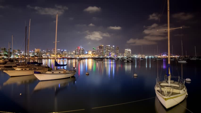 A professional low-angle night cityscape time-lapse capturing a dynamic evening sequence over the San Diego Bay waterfront in San Diego, California. The composition features a beautifully balanced foreground of anchored sailboats and yachts resting on exceptionally calm, glassy water. In the background, the dense corporate skyscraper core of downtown San Diego glows brilliantly, casting long, colorful neon reflections across the bay