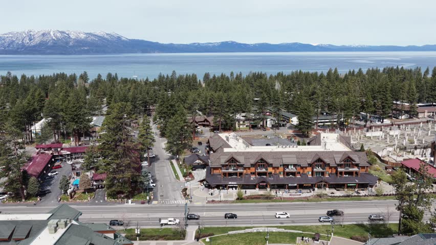 South Lake Tahoe alpine town with rustic lodge-style buildings along main boulevard, dense pine forest and shimmering Lake Tahoe with snow-capped peaks beyond. California, USA.Aerial lateral wide shot