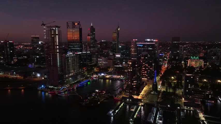 Establishing shot gliding toward Perth CBD at night with illuminated skyline reflected on Swan River waterfront and colorful Elizabeth Quay precinct below. Perth, Aust