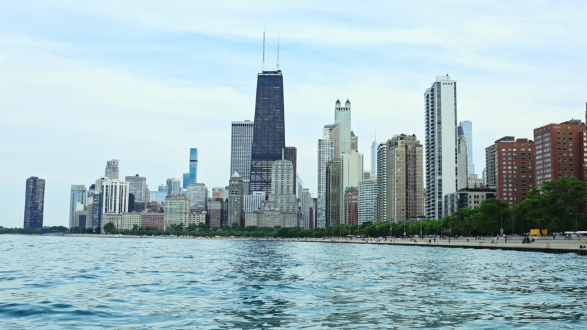 The scene shows the Chicago skyline with many tall buildings next to Lake Michigan. People walk along the waterfront under a cloudy sky. Boats are visible on the lake.