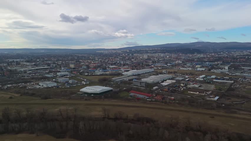 In Miskolc, a wide drone view captures the urban landscape with residential blocks, industrial parks, surrounding hills and nearby trees under an overcast sky.