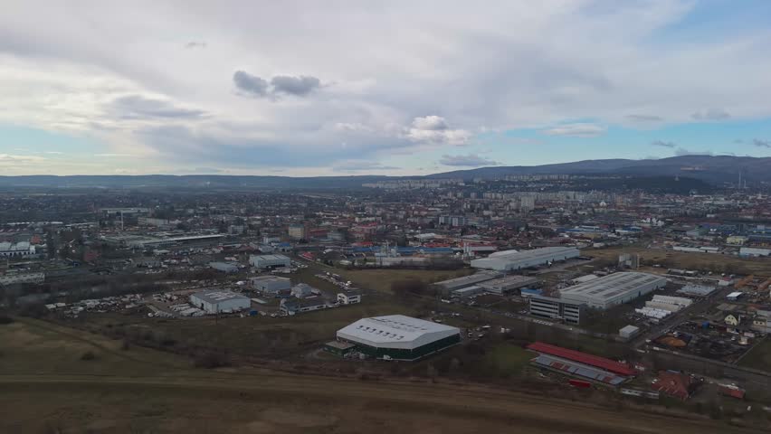 In Miskolc, Hungary, a drone view captures the urban landscape with industrial zones, residential blocks, and surrounding hills and mountains under overcast skies.