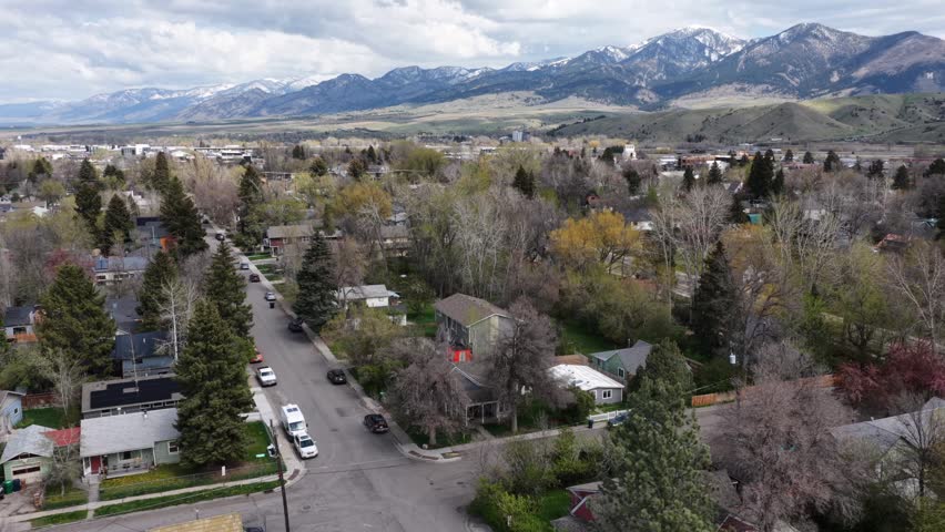 Bozeman tree-lined residential neighborhood with single-family homes, wide valley and snow-capped Bridger Mountains rising steeply beyond under cloudy sky. Montana, USA. Aerial flyover shot.