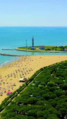 Vertical video. Italy, Jesolo. The Faro di Piave Vecchia lighthouse dominates the river mouth where the Sile River meets the Adriatic Sea, its 48-meter cylindrical tower painted in striking black and white horizontal bands visible for 18 nautical miles. The lighthouse stands on a manicured grassy peninsula enclosed by rocky breakwaters, with a three-story harbor control building at its base that serves as the Jesolo Coast Guard headquarters. Originally built in 1846 under Austrian rule for military and navigational purposes, the structure was destroyed by German forces during World War II and faithfully reconstructed between 1949 and 1951 by the Civil Engineers of Venice. A spiral staircase of 243 Istrian stone steps leads to the lantern gallery. The surrounding scene captures the contrast between the deep turquoise Adriatic waters and the lighter green river currents, with a wide sandy beach stretching along the coast dotted with colorful umbrellas and sunbathers. Dense green pine forests line the shore behind the beach, while the coastal town extends inland with low-rise buildings and tree-lined avenues. The aerial perspective reveals the full geometry of the twin breakwaters framing the harbor entrance, the orderly rows of beach umbrellas, and the long uninterrupted shoreline reaching toward the horizon under a clear summer sky. Aerial View, Point of interest. Rich colors