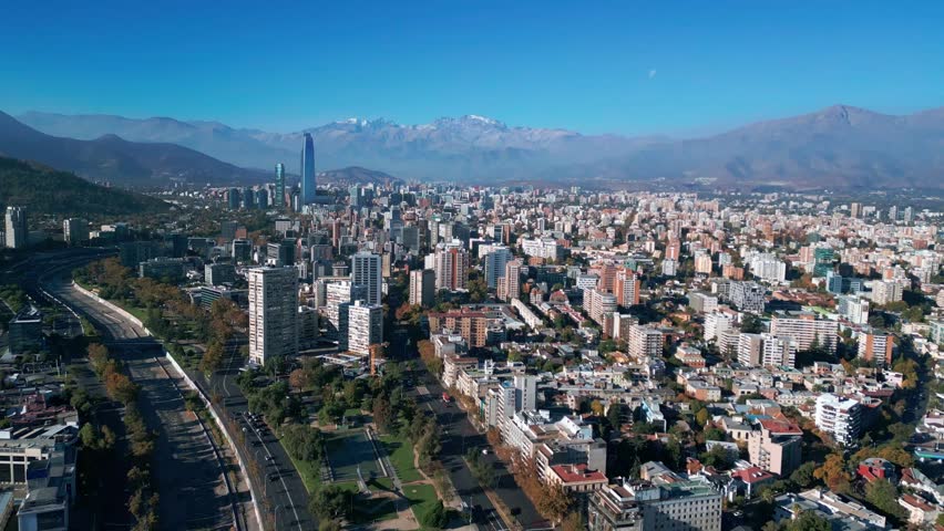 Advancing flight over vast residential urban sprawl with Alameda highway corridor and Costanera Center tower rising against snow-capped Andes. Santiago, Chile