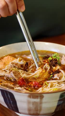 A person uses metal chopsticks to lift rice noodles from a ceramic bowl of soup.