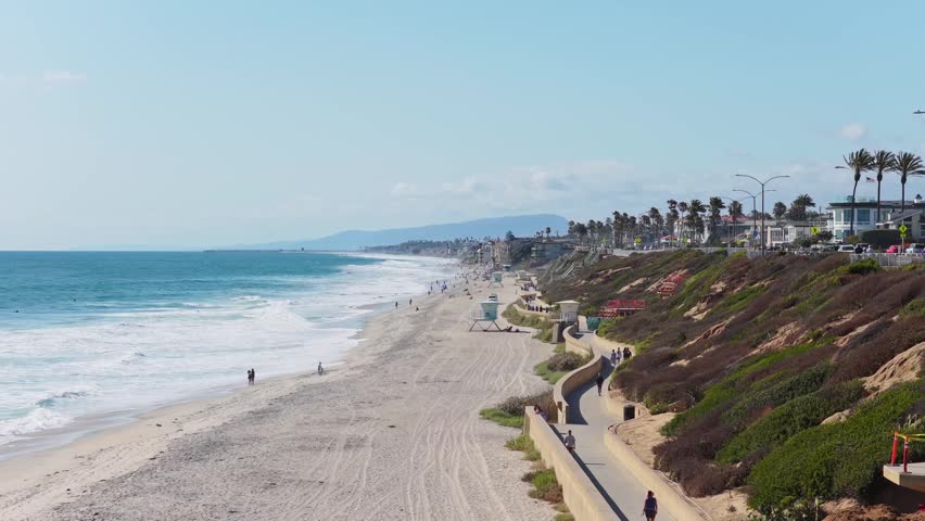 Scenic aerial of Carlsbad State Beach in California showing pedestrians walking on seaside promenade and Pacific Ocean waves crashing on sandy shore
