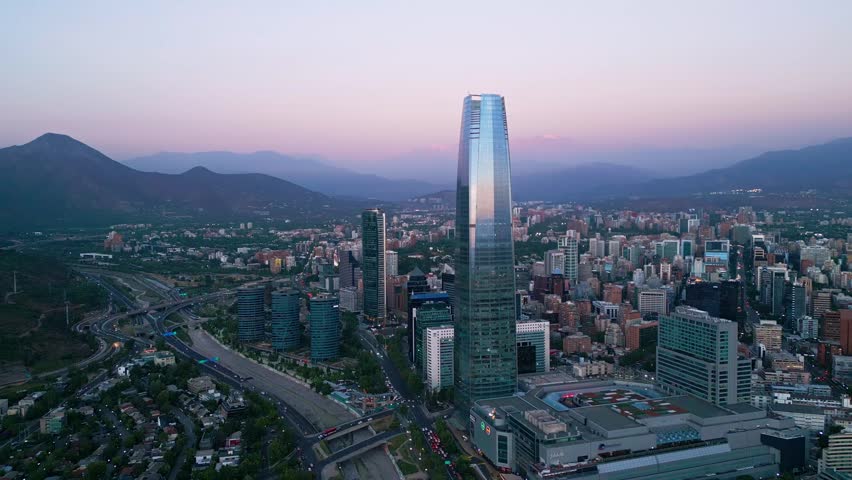 Descending flight toward Costanera Center tower with financial district, Mapocho river interchange and Andes silhouette under pink purple sunset sky. Santiago, Chile