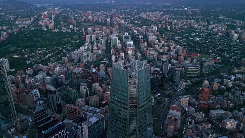 Circling flight over Costanera Center rooftop revealing dense Santiago urban grid, green parks, residential districts and city limits at dusk. Santiago, Chile