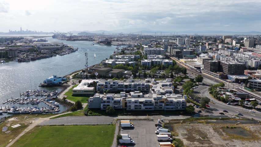 Oakland waterfront with marina, residential blocks and Port of Oakland cranes along estuary, San Francisco skyline faint in distance under partly cloudy sky. California, USA. Aerial view.