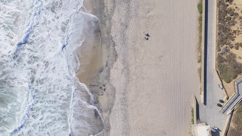 Top down scenery capturing Carlsbad, California, ocean waves crash onto shore, pedestrians enjoy sandy expanse, vehicles move along coastal highway
