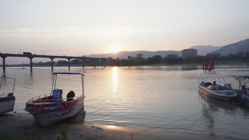 Golden sun dips over calm river waters. Boats rest gently near shore, painted in bright colors. Bridge arches across distant water, connecting land masses. Peaceful glow bathes scene