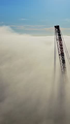 Construction crane emerging above dense clouds against blue sky, drone vertical silhouette