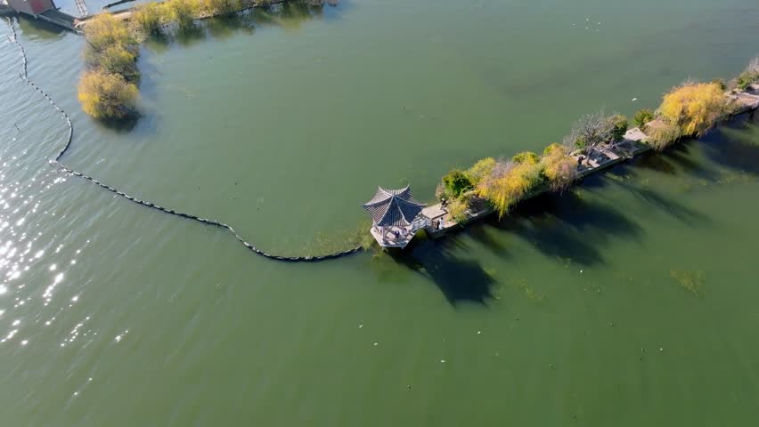 An aerial perspective captures a traditional pavilion surrounded by the serene waters of Erhai Lake in Dali, Yunnan. The scene showcases autumn foliage and a winding path leading to the structure, evoking tranquility.