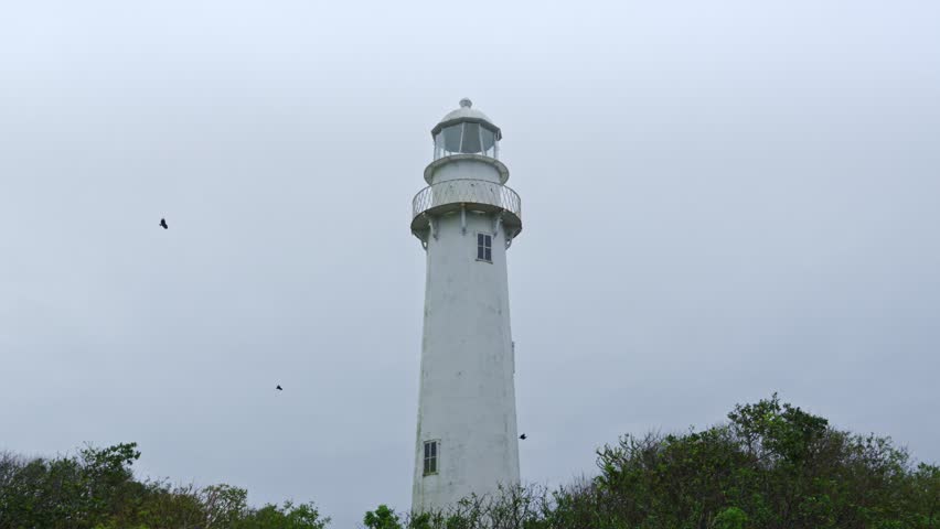 White lighthouse stand tall on an island under a dramatic cloudy sky, birds fly around. Use for travel