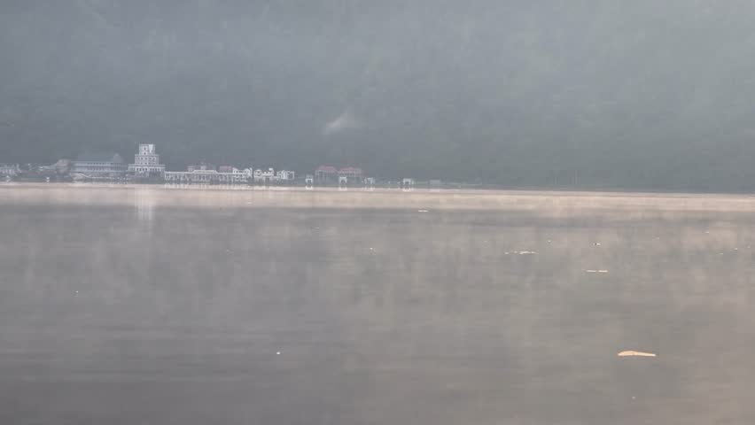 Misty lake surface with moving fog water haze at sunrise morning view of peaceful lake water with distant houses and mountain background.