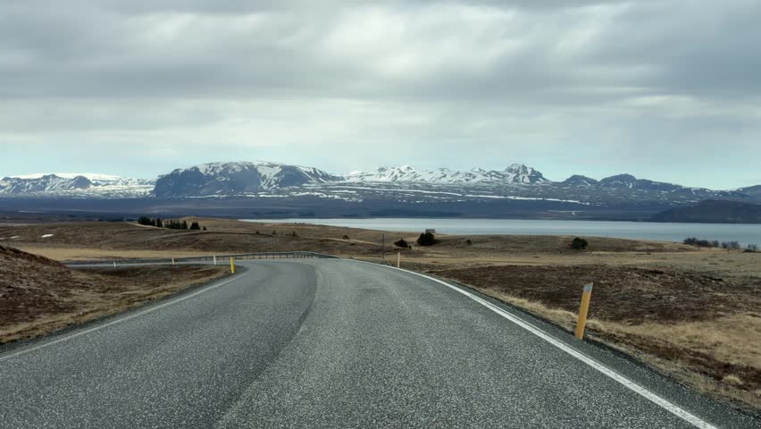 Iceland. Scenic view of a winding road leading through a mountainous landscape with snow-capped peaks and a lake visible in the distance under a cloudy sky